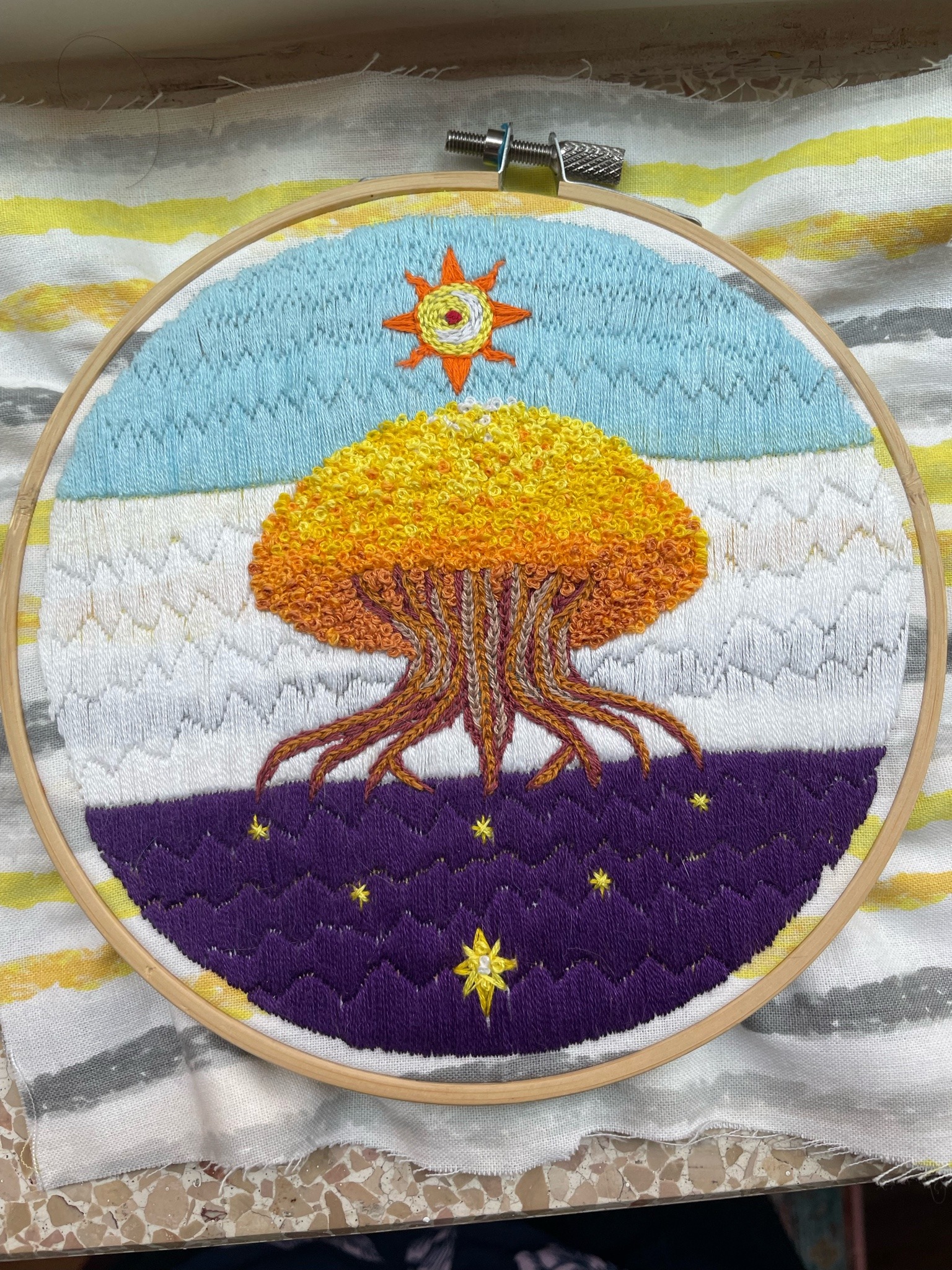 Embroidery piece in the hoop of the Crest of Whitestone from campaign one of Critcal Role. The Sun Tree, a large tree with golden leaves and exposed roots, stands on a field of blue, white, and purple. A stylized sun hangs above the tree, and contains a small white crescent moon, Catha, and a small red dot of a moon, Ruidus. Six stars hang under the tree's roots.